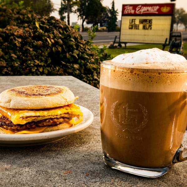 Breakfast sandwich and foamy coffee mug on an outdoor table, with a Tito's Espresso sign.