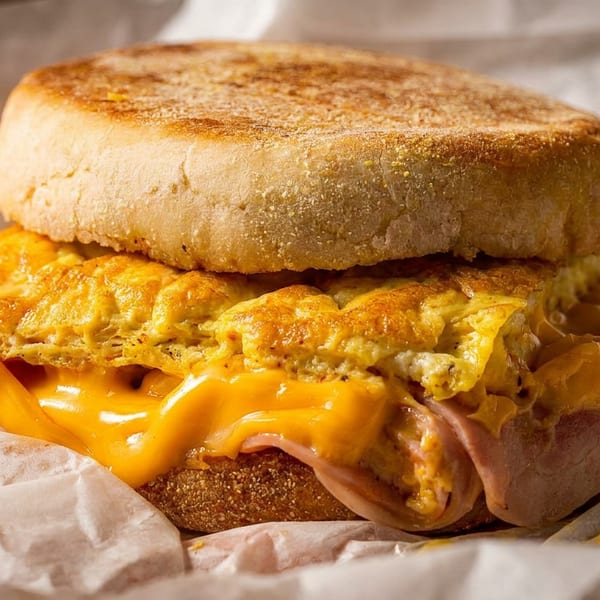 Breakfast sandwich with egg, melted cheese, and ham on an English muffin, wrapped in paper.