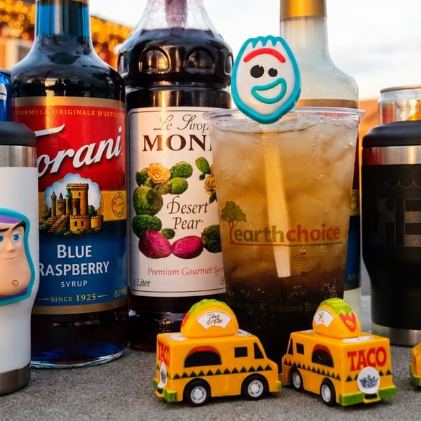 Various flavored syrups, iced drinks with Toy Story toppers, tumblers, and toy taco trucks outdoors.
